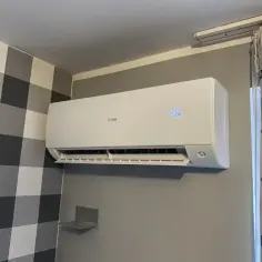 Biała klimatyzacja Haier zamontowana na szarej ścianie, obok wzorzystej tapety i zasłon.