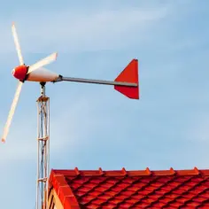Mała turbina wiatrowa na maszcie nad czerwonym dachem, na tle błękitnego nieba.