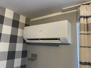 Biała klimatyzacja Haier zamontowana na szarej ścianie, obok wzorzystej tapety i zasłon.